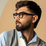 Ares Black Clear Square Sunglasses – Sunglassic.com. Male model in bold black square-frame sunglasses with clear lenses, styled in a smart blue shirt and knitted vest, posing thoughtfully against a soft beige background.